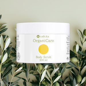 CaliVita OrganiCare Body Scrub 200 ml u bijeloj posudici s žutim krugom okružen svježim maslinovim grančicama na prirodnoj pozadini